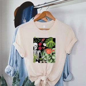 May include: A beige t-shirt with a graphic print of a skeleton holding a watering can surrounded by plants and flowers. The text on the watering can reads "You make me feel alive".
