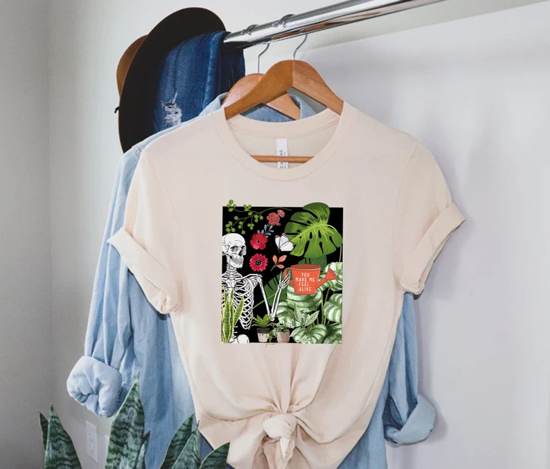 May include: A beige t-shirt with a graphic print of a skeleton holding a watering can surrounded by plants and flowers. The text on the watering can reads "You make me feel alive".