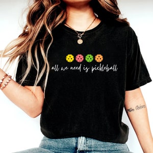 May include: A black t-shirt with a white graphic that says "All we need is pickleball" and four colorful pickleball paddles.