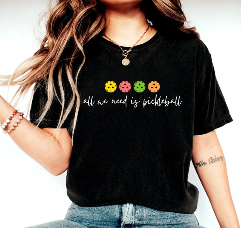May include: A black t-shirt with a white graphic that says "All we need is pickleball" and four colorful pickleball paddles.