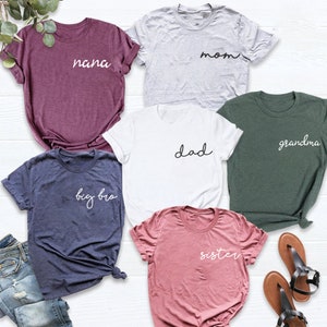 Peut inclure: Six t-shirts avec des textes diff&eacute;rents sur chacun. Les t-shirts sont de diff&eacute;rentes couleurs, notamment violet, gris, blanc, vert, bleu et rose. Le texte sur les t-shirts comprend "nana", "mom", "dad", "grandma", "big bro" et "sister".