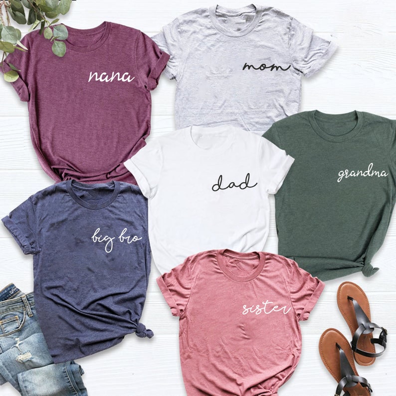 Peut inclure: Six t-shirts avec des textes diff&eacute;rents sur chacun. Les t-shirts sont de diff&eacute;rentes couleurs, notamment violet, gris, blanc, vert, bleu et rose. Le texte sur les t-shirts comprend "nana", "mom", "dad", "grandma", "big bro" et "sister".