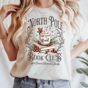K&ouml;nnte beinhalten: Ein wei&szlig;es T-Shirt mit einem grafischen Design einer Tasse hei&szlig;er Schokolade mit einem Lebkuchenmann und einem Zuckerstangen, umgeben von B&uuml;chern und Gr&uuml;n. Der Text lautet "North Pole Book Club Hot Cocoa Served Daily".