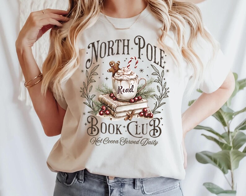 K&ouml;nnte beinhalten: Ein wei&szlig;es T-Shirt mit einem grafischen Design einer Tasse hei&szlig;er Schokolade mit einem Lebkuchenmann und einem Zuckerstangen, umgeben von B&uuml;chern und Gr&uuml;n. Der Text lautet "North Pole Book Club Hot Cocoa Served Daily".