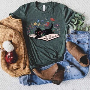 May include: A dark green t-shirt with a black cat reading a book surrounded by colorful flowers. The cat is sitting on the open book.