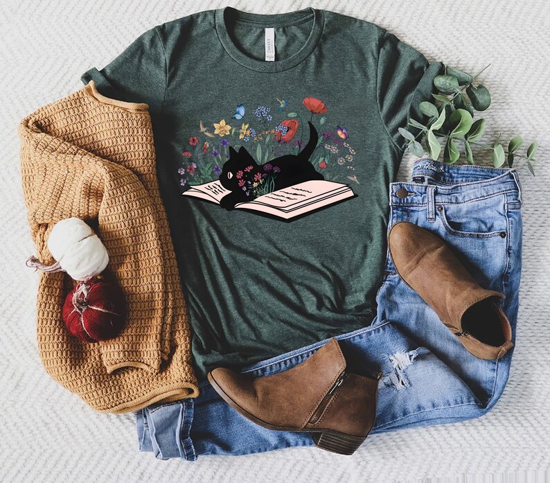 May include: A dark green t-shirt with a black cat reading a book surrounded by colorful flowers. The cat is sitting on the open book.
