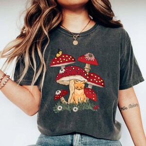 May include: A dark gray t-shirt with a graphic of a cat sitting under a cluster of red and white mushrooms. The mushrooms have white spots and a butterfly is perched on one of the mushrooms.
