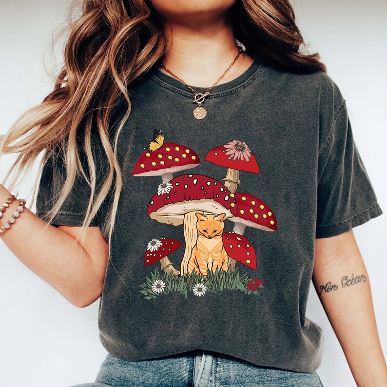 May include: A dark gray t-shirt with a graphic of a cat sitting under a cluster of red and white mushrooms. The mushrooms have white spots and a butterfly is perched on one of the mushrooms.