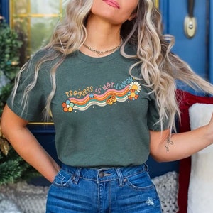 K&ouml;nnte beinhalten: Ein gr&uuml;nes T-Shirt mit dem Text "Progress is not linear" in Regenbogen-Schrift mit Blumen und Sternen. Das Shirt wird von einer Person mit langen blonden Haaren getragen.