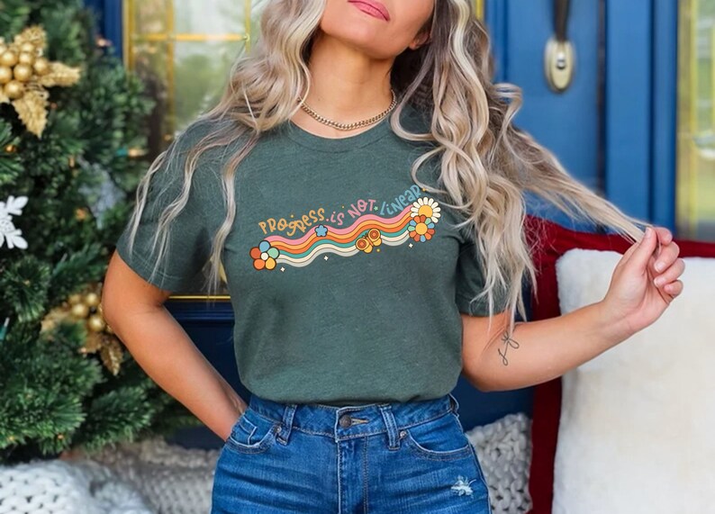 K&ouml;nnte beinhalten: Ein gr&uuml;nes T-Shirt mit dem Text "Progress is not linear" in Regenbogen-Schrift mit Blumen und Sternen. Das Shirt wird von einer Person mit langen blonden Haaren getragen.