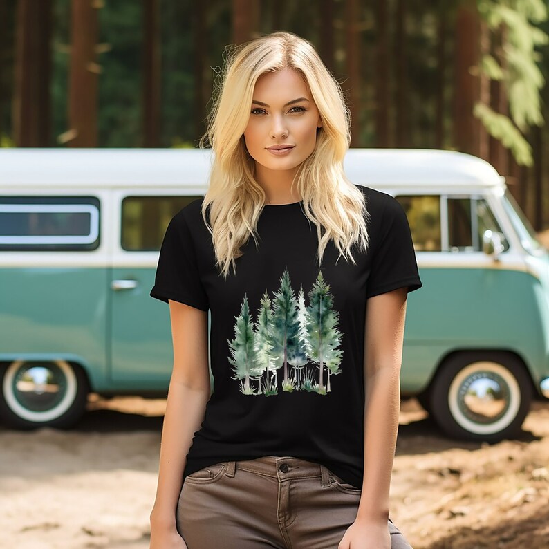 Puede incluir: Una camiseta negra con un dise&ntilde;o de acuarela de un bosque con &aacute;rboles verdes. La camiseta la lleva puesta una persona con el pelo largo y rubio. La persona est&aacute; de pie delante de una furgoneta vintage.