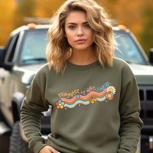 K&ouml;nnte beinhalten: Eine Frau tr&auml;gt ein gr&uuml;nes Sweatshirt mit einem Regenbogen- und Blumenmotiv, auf dem "Progress is not linear" steht.