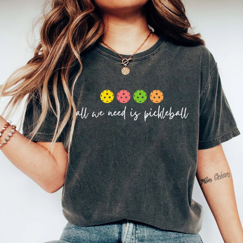 May include: A dark gray t-shirt with a white graphic that says "all we need is pickleball" and features five colorful pickleball paddles.