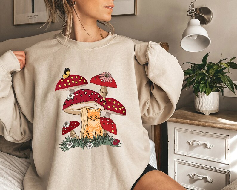 May include: A beige sweatshirt with a graphic of a cat sitting under a cluster of red and white mushrooms with a butterfly on one of the mushrooms.