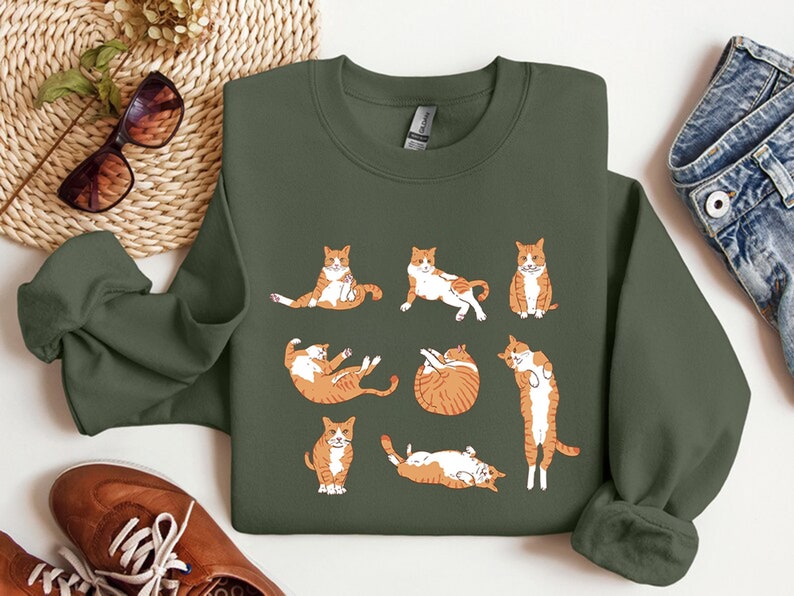 K&ouml;nnte beinhalten: Ein gr&uuml;ner Pullover mit einem grafischen Aufdruck von orangefarbenen und wei&szlig;en Katzen in verschiedenen Yoga-Posen.