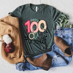 May include: A green t-shirt with the text "Happy 100th day of school" in colorful letters. The shirt is laid out on a white surface with a brown sweater, blue jeans, and brown boots.