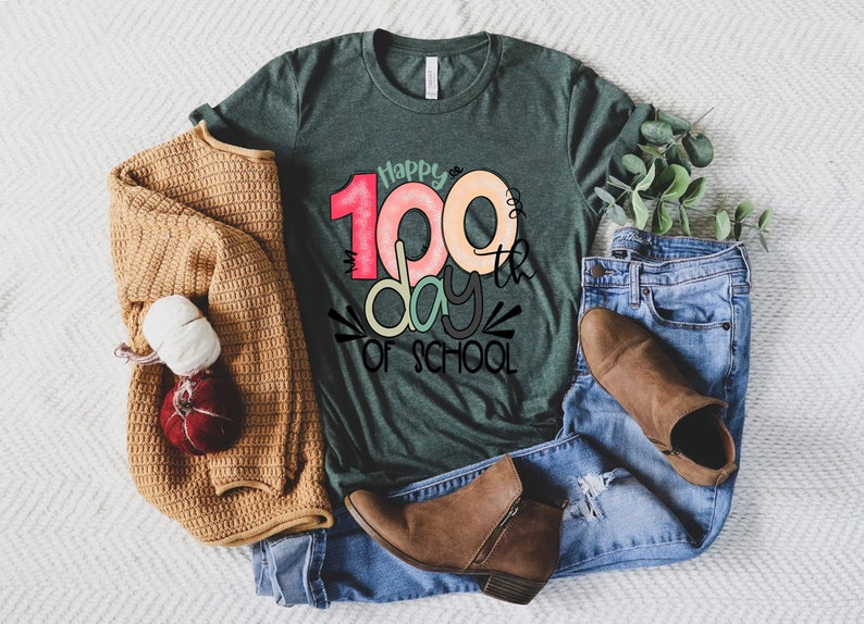 May include: A green t-shirt with the text "Happy 100th day of school" in colorful letters. The shirt is laid out on a white surface with a brown sweater, blue jeans, and brown boots.