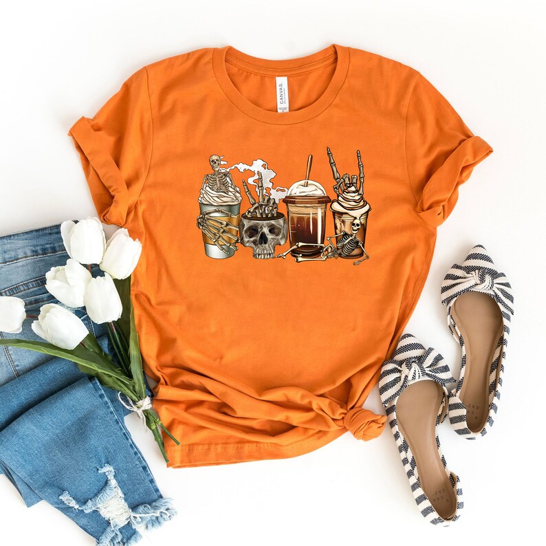 K&ouml;nnte beinhalten: Orangefarbenes T-Shirt mit einem Motiv von vier Kaffeetassen mit Skeletten und Sch&auml;deln. Die Kaffeetassen haben Schlagsahne und verschiedene Toppings.
