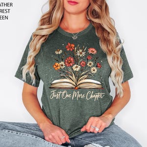 以下が含まれることがあります： ヘザーフォレストグリーンのTシャツに、花柄のデザインと「Just One More Chapter」という文字が書かれた開いた本がプリントされています。