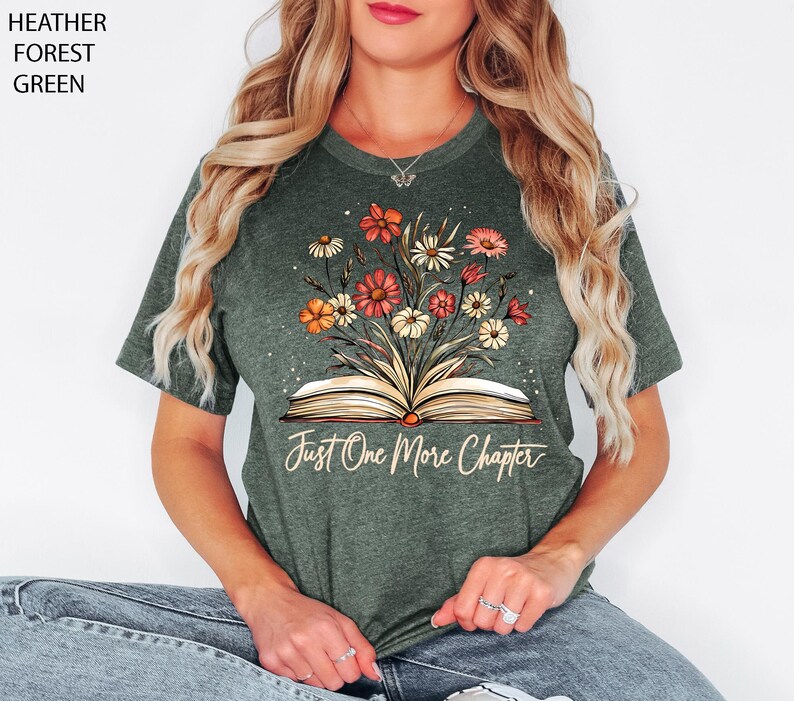 以下が含まれることがあります： ヘザーフォレストグリーンのTシャツに、花柄のデザインと「Just One More Chapter」という文字が書かれた開いた本がプリントされています。