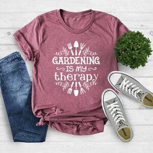 May include: A maroon t-shirt with white text that reads "Gardening is my therapy" and gardening tools and leaves around the text.