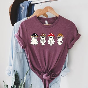 May include: A maroon t-shirt featuring a graphic of five cartoon mushrooms dressed as ghosts. The mushrooms are red, brown and black with white spots.