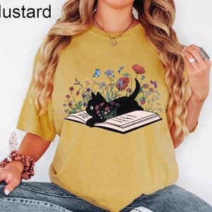 May include: A mustard yellow t-shirt with a black cat reading a book surrounded by flowers and butterflies. The text "Mustard" is in the top left corner of the image.