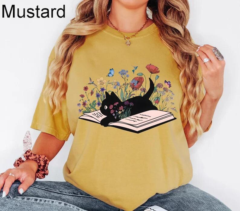 May include: A mustard yellow t-shirt with a black cat reading a book surrounded by flowers and butterflies. The text "Mustard" is in the top left corner of the image.