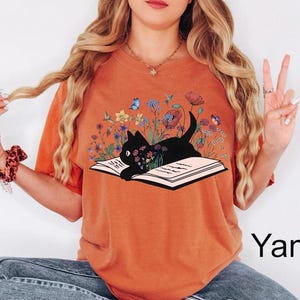 May include: A burnt orange t-shirt with a black cat reading a book surrounded by flowers and butterflies. The text "Yam" is visible in the bottom right corner of the image.