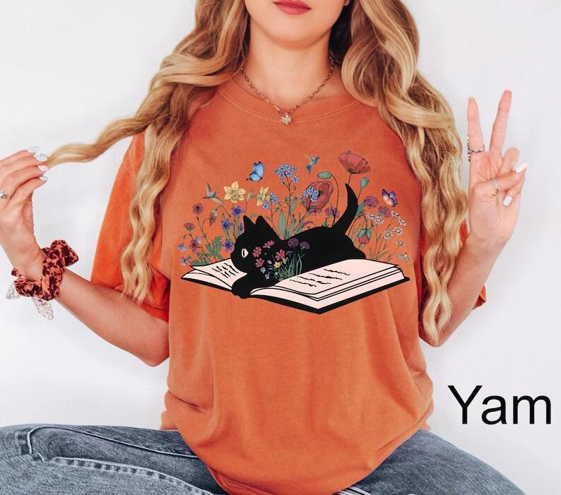 May include: A burnt orange t-shirt with a black cat reading a book surrounded by flowers and butterflies. The text "Yam" is visible in the bottom right corner of the image.