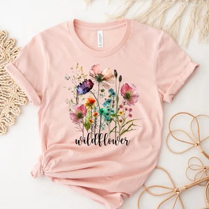 May include: A pink t-shirt featuring a watercolour floral design and the word "wildflower" printed in black.