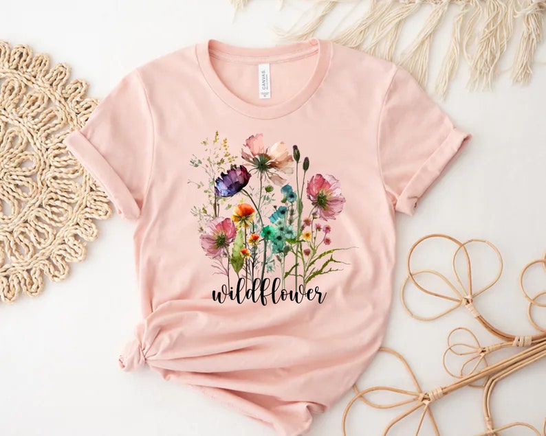 May include: A pink t-shirt featuring a watercolour floral design and the word "wildflower" printed in black.
