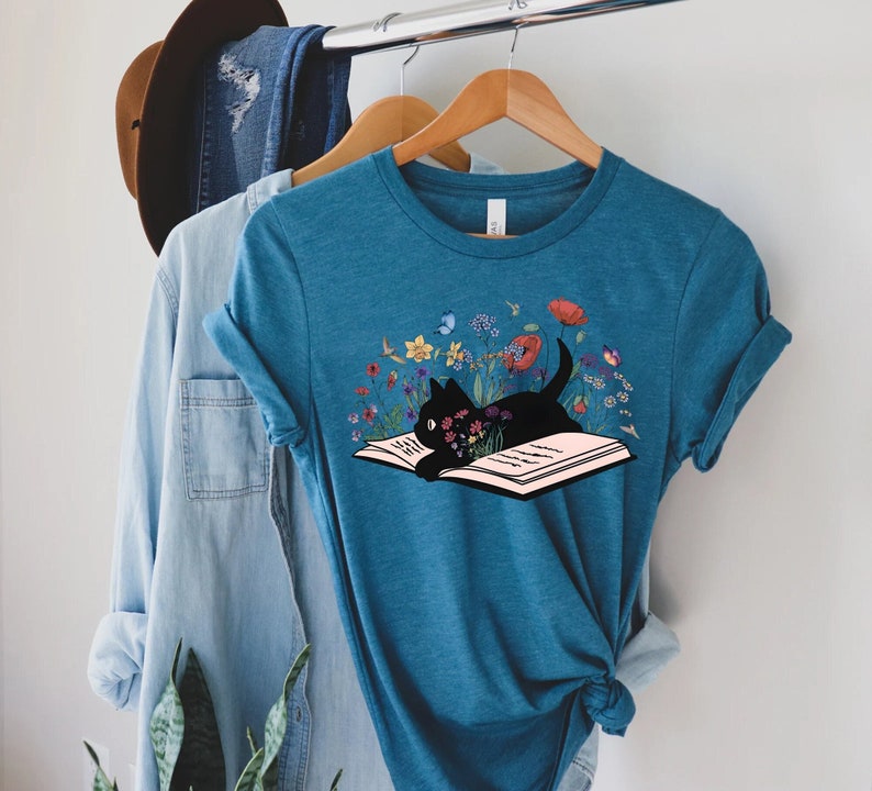 May include: A teal blue t-shirt with a black cat reading a book surrounded by colorful flowers. The cat is lying on the open book with its head resting on the pages.