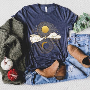 May include: A navy blue t-shirt with a gold sun and moon design with clouds and stars. The shirt is laying on a white fuzzy surface with a pair of blue jeans, a brown sweater, and brown boots.