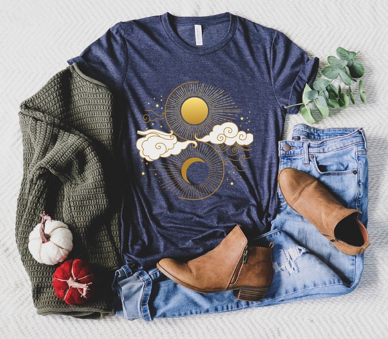 May include: A navy blue t-shirt with a gold sun and moon design with clouds and stars. The shirt is laying on a white fuzzy surface with a pair of blue jeans, a brown sweater, and brown boots.