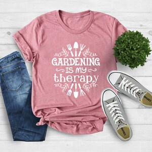 May include: A pink t-shirt with the text "Gardening is my therapy" in white. The text is surrounded by a design of gardening tools and leaves. The shirt is laid out on a wooden surface with a pair of grey trainers and a potted plant.