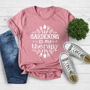 May include: A pink t-shirt with the text "Gardening is my therapy" in white. The text is surrounded by a design of gardening tools and leaves. The shirt is laid out on a wooden surface with a pair of grey trainers and a potted plant.