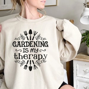 May include: A cream-coloured sweatshirt with black text that reads "Gardening is my therapy" and a design of gardening tools and leaves.