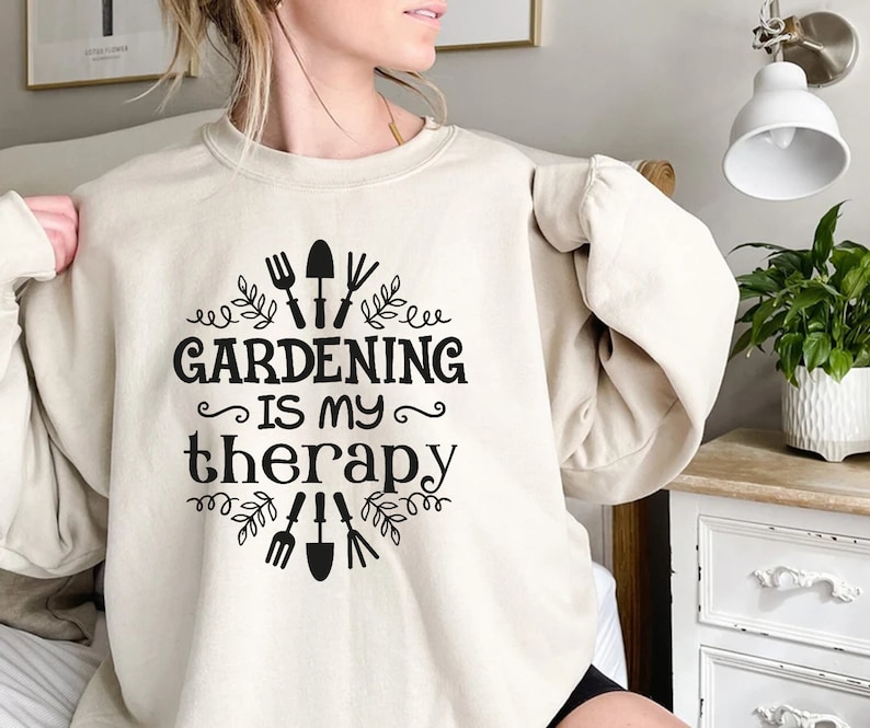 May include: A cream-coloured sweatshirt with black text that reads "Gardening is my therapy" and a design of gardening tools and leaves.