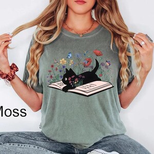 May include: A green t-shirt with a black cat reading a book surrounded by flowers. The cat is sitting on an open book with a floral design. The shirt has the word "Moss" printed on the bottom left corner.
