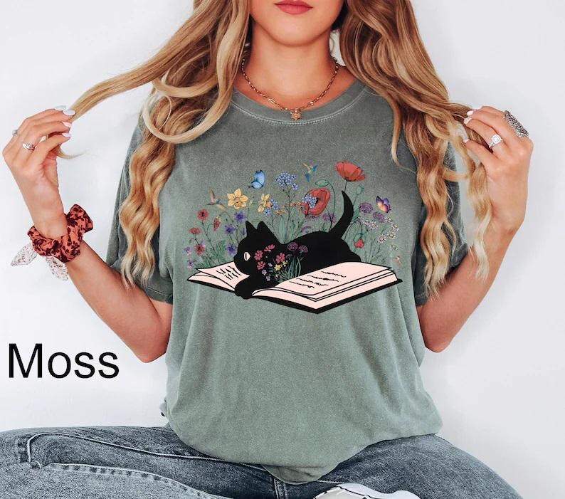 May include: A green t-shirt with a black cat reading a book surrounded by flowers. The cat is sitting on an open book with a floral design. The shirt has the word "Moss" printed on the bottom left corner.