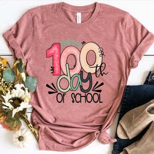 May include: A pink t-shirt with the text "Happy 100th Day of School" in colorful letters and a black and white graphic design.