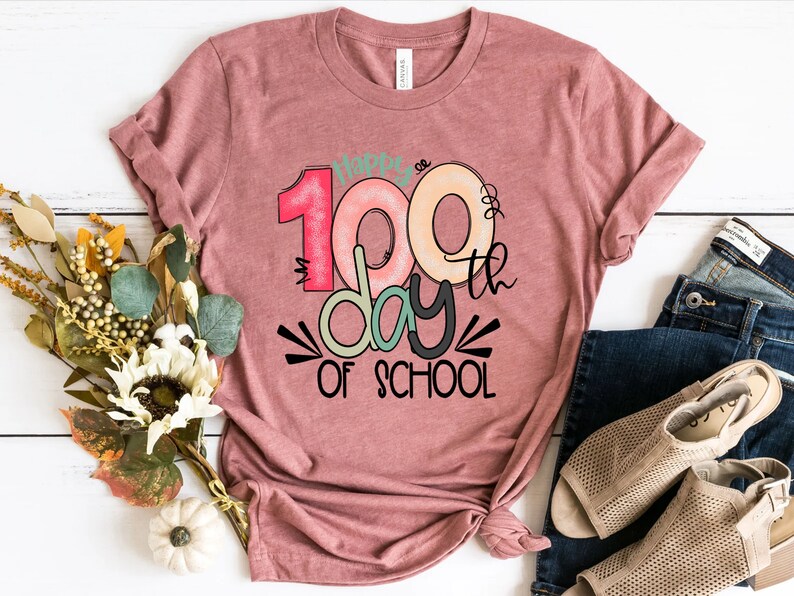 May include: A pink t-shirt with the text "Happy 100th Day of School" in colorful letters and a black and white graphic design.