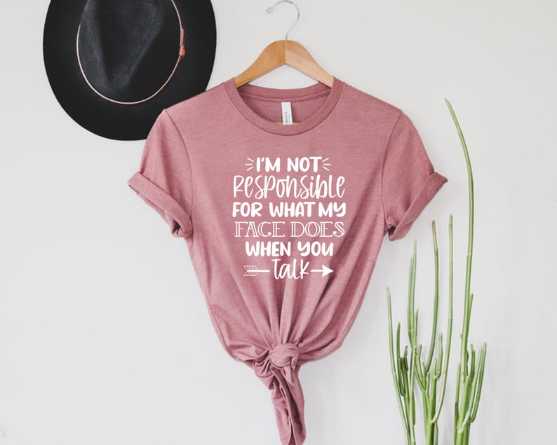 K&ouml;nnte beinhalten: Ein pinkfarbenes T-Shirt mit dem Text "Ich bin nicht verantwortlich f&uuml;r das, was mein Gesicht tut, wenn du sprichst" in wei&szlig;en Buchstaben. Das Shirt hat einen Knoten vorne und wird auf einem Kleiderb&uuml;gel pr&auml;sentiert.