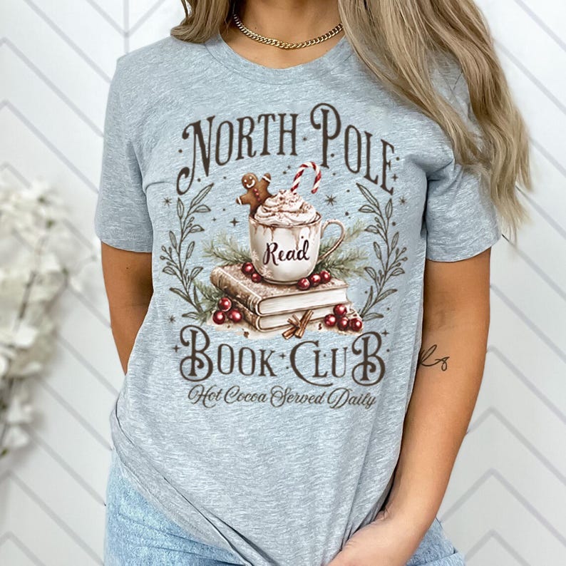 K&ouml;nnte beinhalten: Graues T-Shirt mit einem grafischen Design einer Tasse mit hei&szlig;er Schokolade mit Schlagsahne und einem Lebkuchenmann oben drauf. Die Tasse steht auf einem Stapel B&uuml;cher mit einem Kranz aus Stechpalme und Beeren drumherum. Der Text "North Pole Book Club" befindet sich &uuml;ber der Tasse und "Hot Cocoa Served Daily" unter der Tasse.