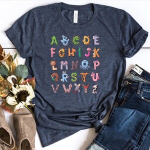 May include: A navy blue t-shirt with a colorful alphabet printed on it. Each letter is decorated with a different animal illustration. The shirt is laid out on a white wooden surface with a pair of brown sandals and blue jeans.