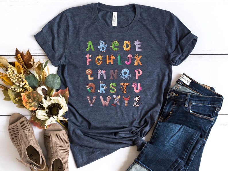 May include: A navy blue t-shirt with a colorful alphabet printed on it. Each letter is decorated with a different animal illustration. The shirt is laid out on a white wooden surface with a pair of brown sandals and blue jeans.