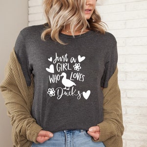 May include: Dark gray t-shirt with the text "Just a GIRL WHO LOVES Ducks" in white. The design includes hearts, a flower, and a duck illustration. The shirt is a casual wear item.