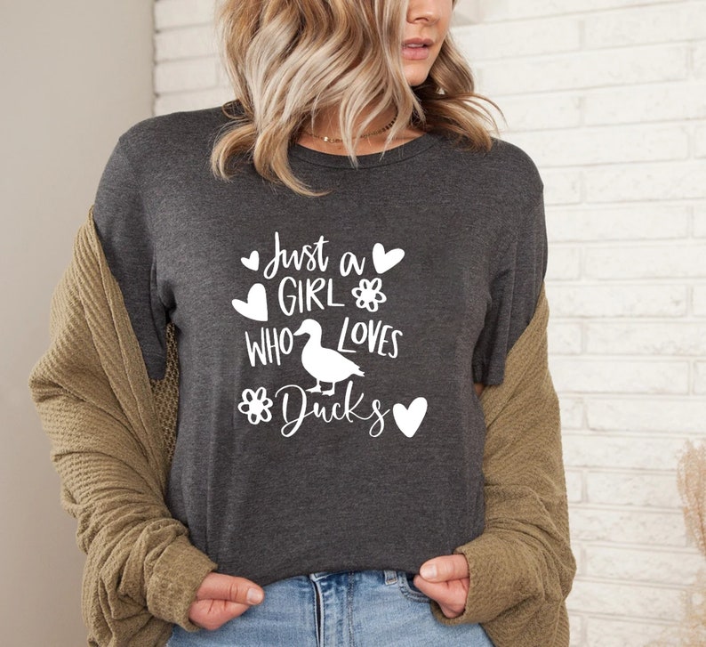 May include: Dark gray t-shirt with the text "Just a GIRL WHO LOVES Ducks" in white. The design includes hearts, a flower, and a duck illustration. The shirt is a casual wear item.
