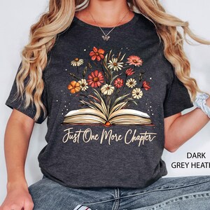 以下が含まれることがあります： ダークグレーヘザーのTシャツに、花柄のデザインと「Just One More Chapter」という文字が書かれた開いた本が描かれています。